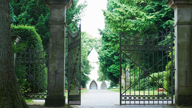 Eisenzaun mit geöffneten Toren, Blick auf einen Weg, der zu einer weißen Kapelle zwischen grünen Bäumen führt.