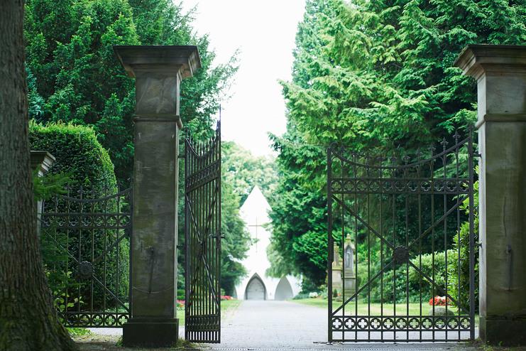Eisenzaun mit geöffneten Toren, Blick auf einen Weg, der zu einer weißen Kapelle zwischen grünen Bäumen führt.