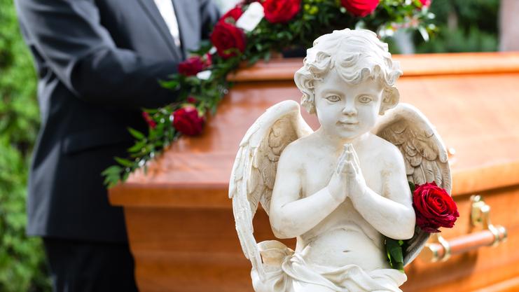 Engelstatue mit gefalteten Händen und einer Rose, im Hintergrund ein mit Blumen geschmückter Sarg.