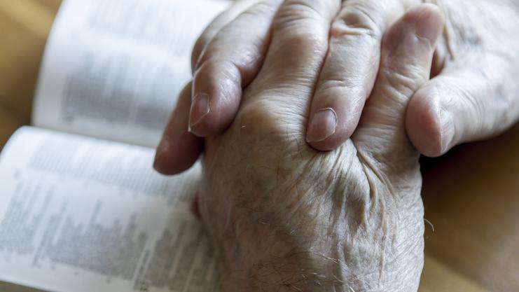 Hände eines älteren Menschen liegen faltig und übereinander auf einem offenen Buch mit Text.