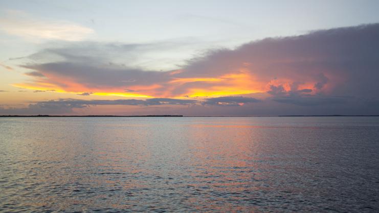 Blick auf einen ruhigen See mit orangefarbenem Sonnenuntergang und sanften Wolken am Horizont.
