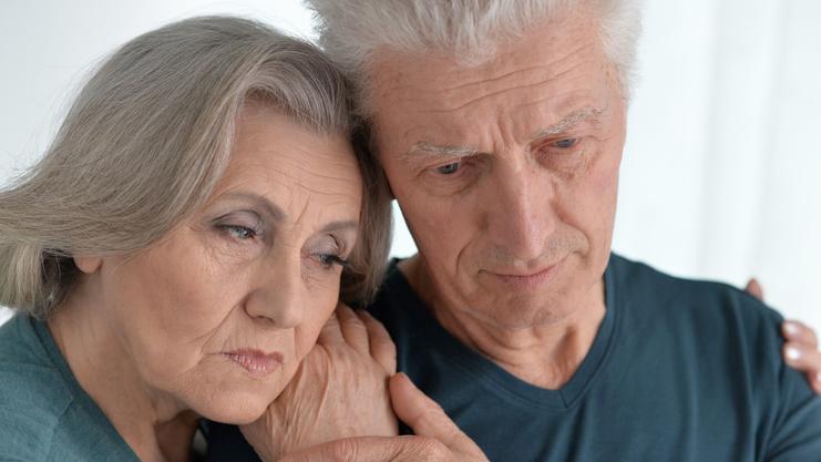 Ein älteres Paar steht eng umschlungen, beide blicken nachdenklich und wirken emotional berührt.